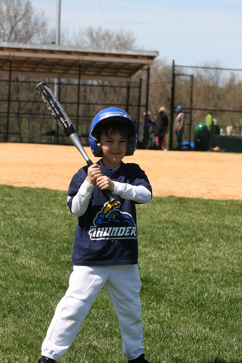 Joey Edwards in a blue "Thunder" baseball shirt and helmet holds a bat, standing on a grassy field with a dirt baseball diamond behind him.