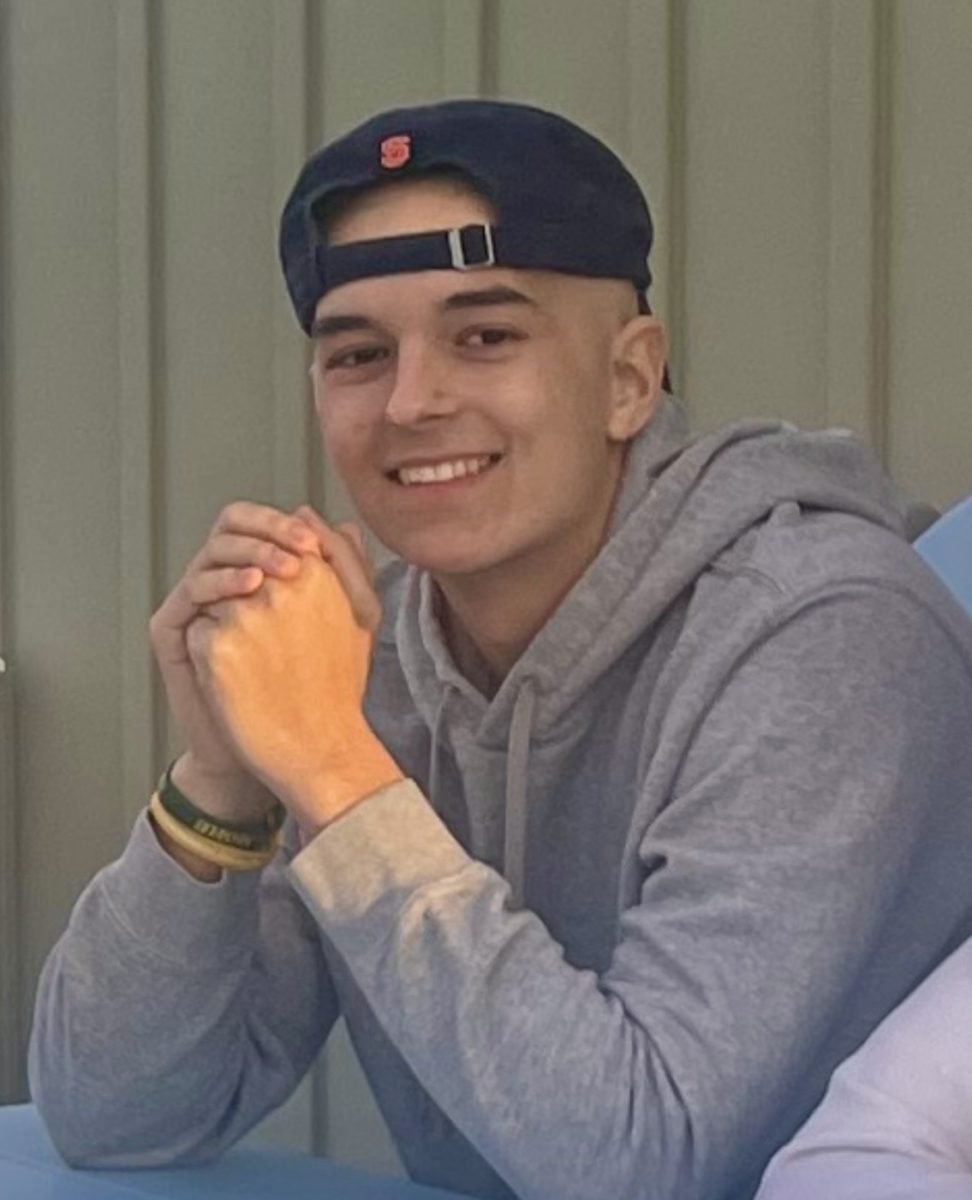 Joey Edwards wearing a backwards cap, gray hoodie, and wristbands, sitting with hands clasped and smiling at the camera.