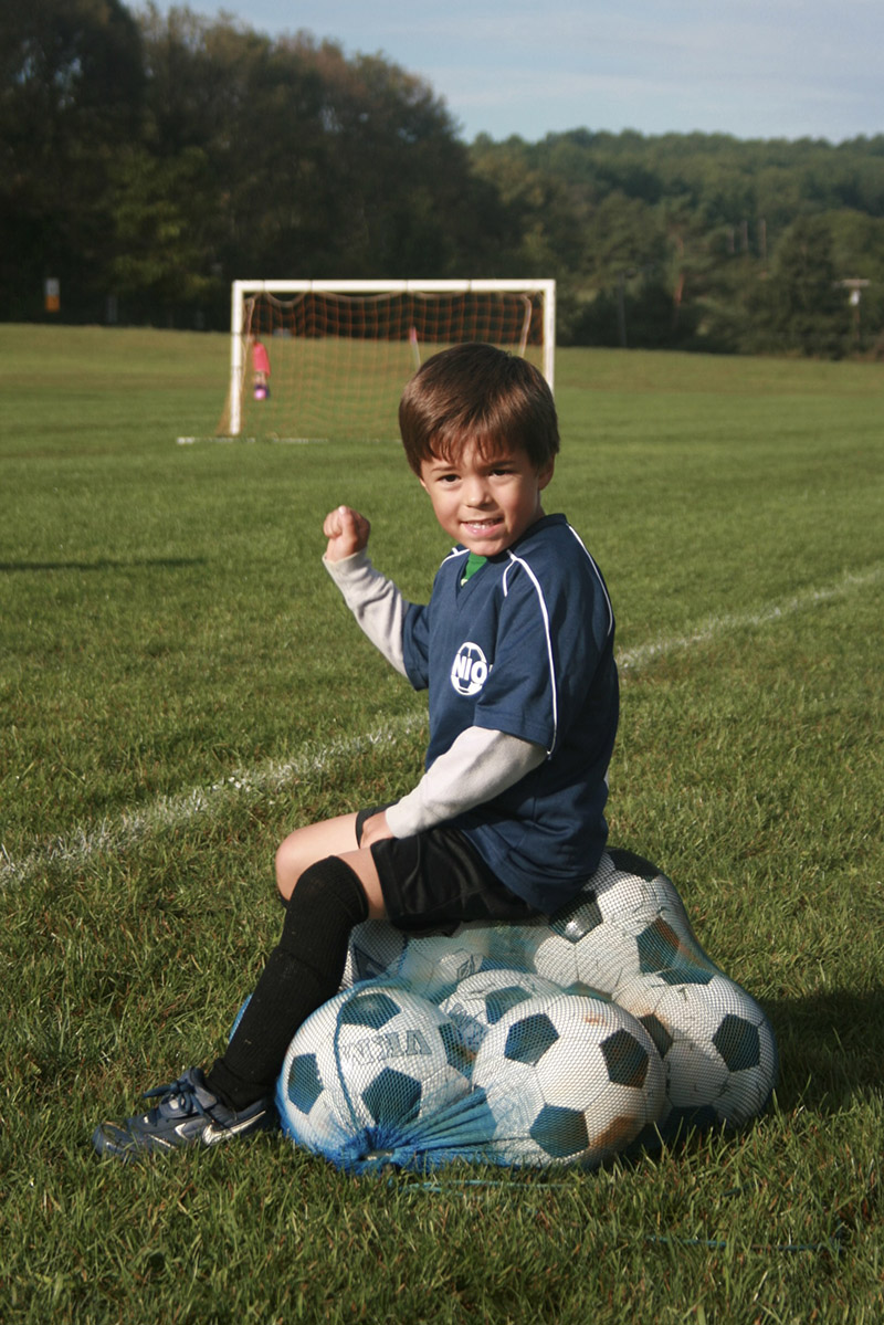 Joey Edwards wearing a soccer uniform sits on a net full of soccer balls on a grass field, raising his fist. A soccer goal and another player are visible in the background.