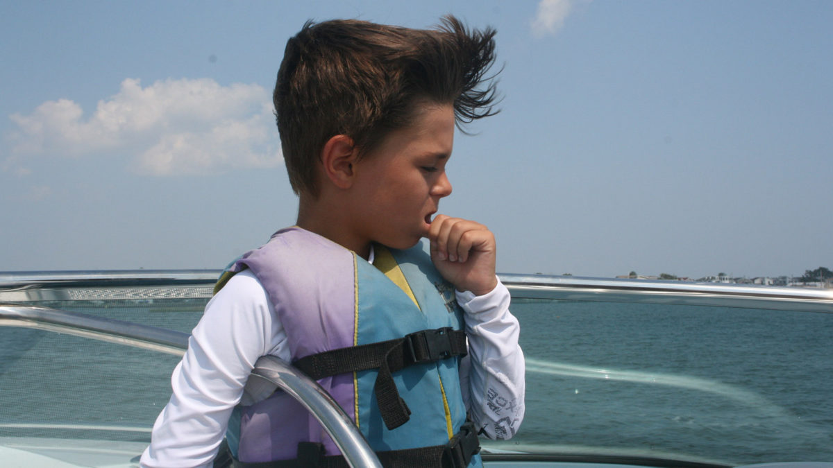 Joey Edwards wearing a life jacket stands on a boat, looking thoughtful with his hand near his mouth. The sea and a clear sky with some clouds are in the background.